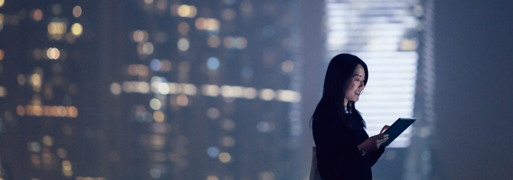Geschäftsfrau, die an einem digitalen Tablet vor einem beleuchteten Hochhaus in der Nacht steht.