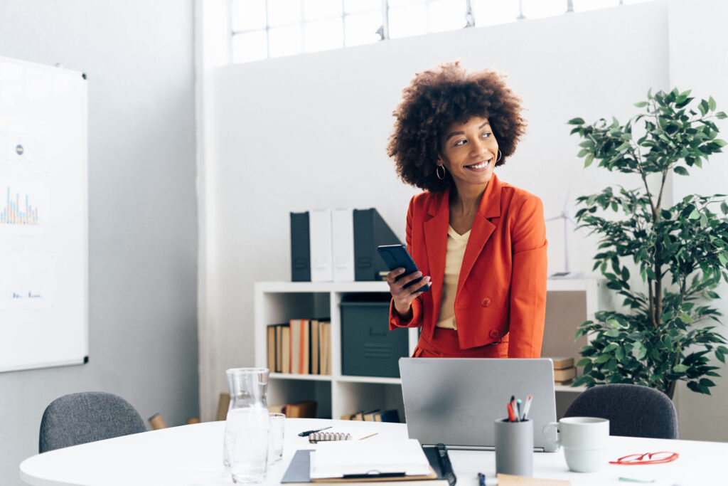 Happy businesswoman holding smart phone standing with laptop at desk in office