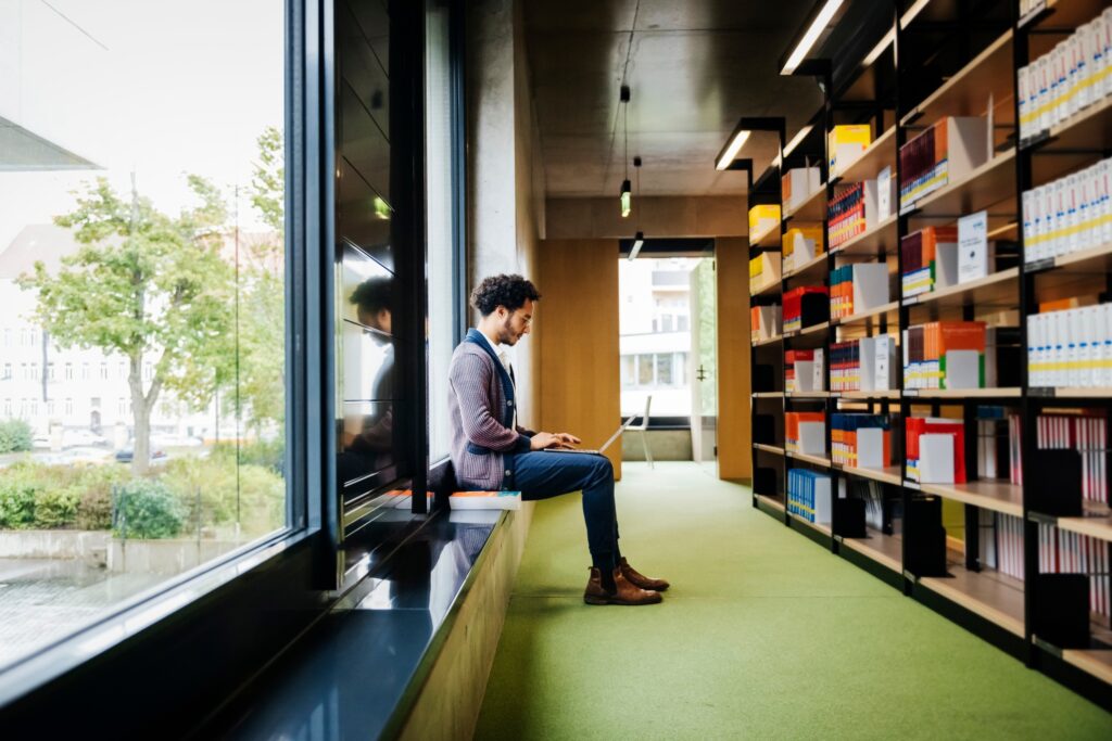 A man sitting by a large window in a modern library space while working on a laptop.