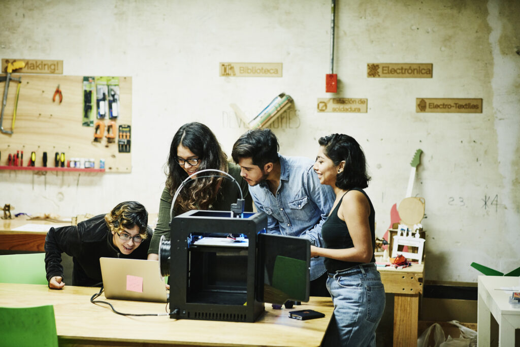 Engineers examining plans on laptop while prototyping parts for project on 3D printer in workshop