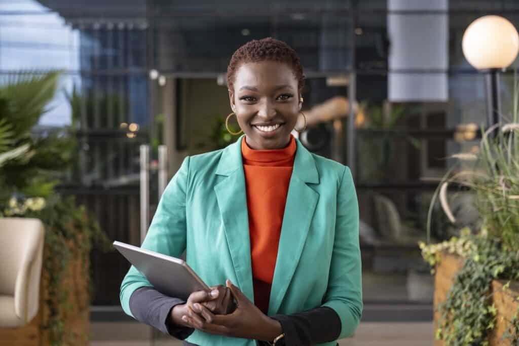 Waist-up view of well-dressed young woman standing in open-air office building lounge, holding laptop, and smiling at camera.