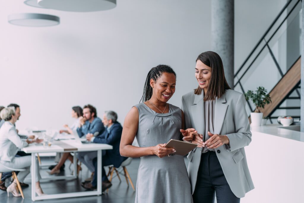 Shot of two female coworkers having a discussion in modern office. Businesswomen in meeting using digital tablet and discussing business strategy. Confident business people working together in the office. Corporate business persons discussing new project and sharing ideas in the workplace. Successful businesswomen standing with their colleagues working in background.