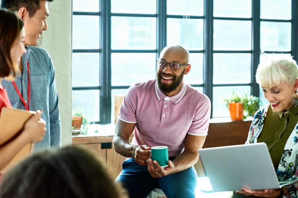 Mid adult African man in glasses with coffee, smiling and talking to colleague, senior woman using laptop and making notes