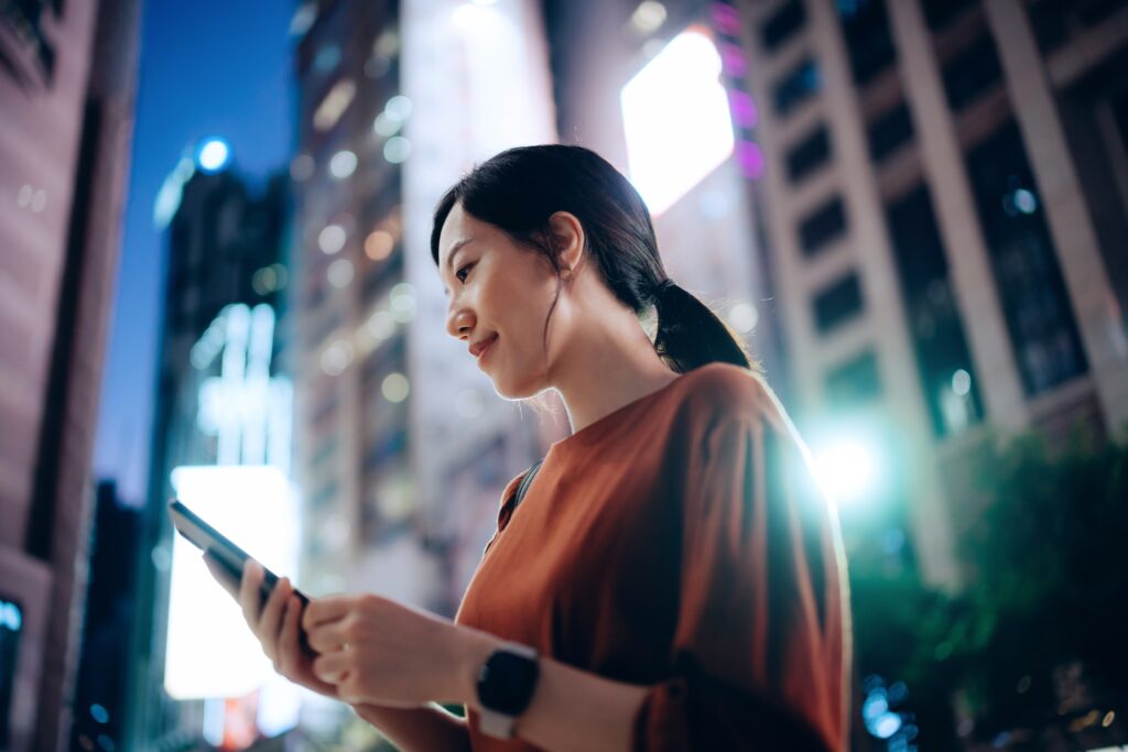 Low angle portrait of confident and beautiful young Asian businesswoman using smartphone while commuting in downtown city street, standing against illuminated urban skyscrapers in the city. Business on the go