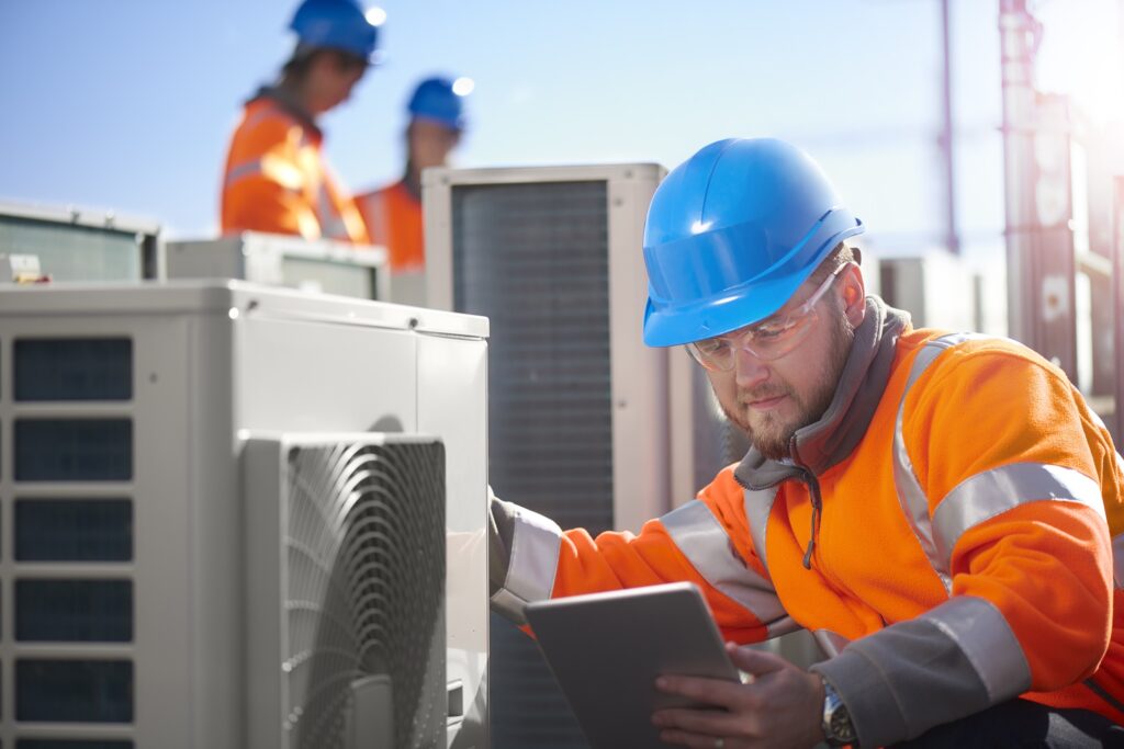 An air conditioning engineer is finishing the installation of several units on a rooftop. Two colleagues can be seen also installing units in the background. They are wearing hi vis jackets, hard hats and safety goggles.