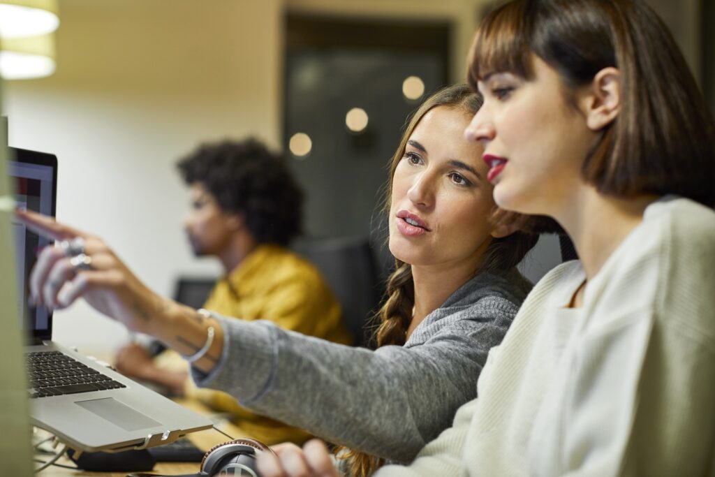 Businesswoman explaining coworker over computer at desk. Female business professionals are discussing in office. They are in smart casuals.