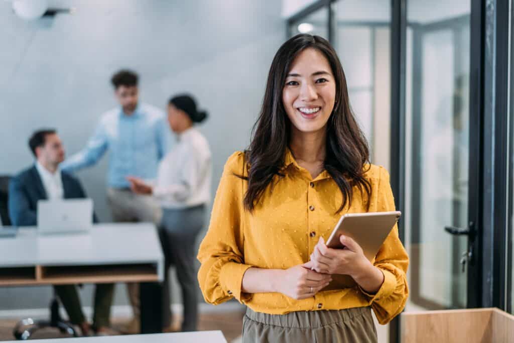 Shot of a beautiful smiling businesswoman standing in front of her team and holding digital tablet. Portrait of successful businesswoman standing with her colleagues working in background.